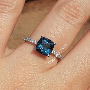 May include: A silver ring featuring a large, square-shaped blue topaz gemstone surrounded by smaller, clear gemstones. The ring is worn on a finger.