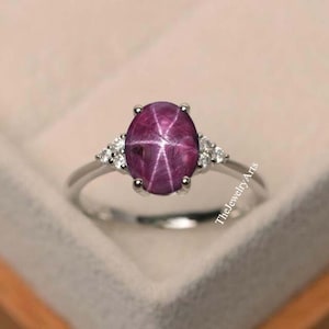 May include: A silver ring featuring a large oval star ruby gemstone, surrounded by smaller diamonds. The ruby is a deep purple colour with a star-like pattern.