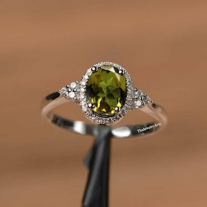 Può includere: Un anello d'argento con una gemma ovale verde oliva, circondata da un alone di piccole pietre trasparenti. Ulteriori pietre trasparenti sono incastonate su entrambi i lati della pietra principale. L'anello è presentato su uno sfondo neutro.