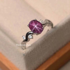 K&ouml;nnte beinhalten: Ein silberner Ring mit einem rosafarbenen Sternrubin-Edelstein und kleinen klaren Edelsteinen. Der Ring hat ein zartes, geschwungenes Design.