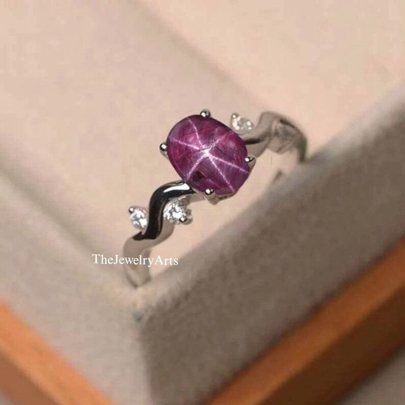 K&ouml;nnte beinhalten: Ein silberner Ring mit einem rosafarbenen Sternrubin-Edelstein und kleinen klaren Edelsteinen. Der Ring hat ein zartes, geschwungenes Design.