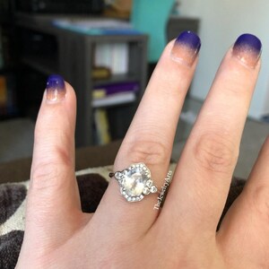 May include: A silver ring with a large, clear, oval-shaped stone, surrounded by smaller stones. The ring is on a hand with ombre nail polish in shades of blue and peach. The Jewelry Arts is written on the side of the hand.