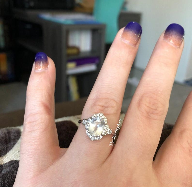 May include: A silver ring with a large, clear, oval-shaped stone, surrounded by smaller stones. The ring is on a hand with ombre nail polish in shades of blue and peach. The Jewelry Arts is written on the side of the hand.
