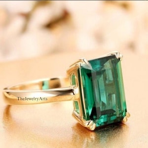May include: A gold ring featuring a large emerald green gemstone set in a square shape. The ring is displayed on a gold surface.