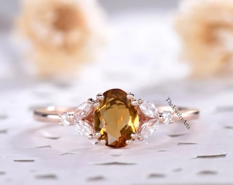 May include: A rose gold ring featuring a central oval-cut yellow citrine gemstone flanked by marquise-cut and round-cut white stones in a prong setting. The text TheJewelryArts is visible on the right side of the ring band.