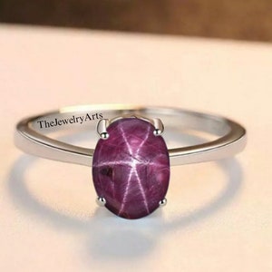 May include: A silver ring featuring a large oval ruby gemstone. The ruby has a star-like pattern within it. The ring is inscribed with the text "TheJewelryArts".