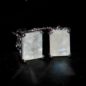 Puede incluir: Un par de pendientes de botón de piedra lunar rectangulares. Los pendientes tienen un engaste plateado y una piedra clara e iridiscente. El texto "TheJewelryArts" es visible en la esquina superior derecha.