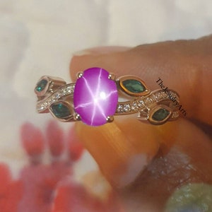 Op de afbeelding: Een zilveren ring met een roze ster-saffier edelsteen, omringd door kleinere groene edelstenen en witte diamanten. De ring heeft een delicaat bladontwerp.