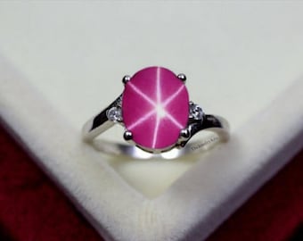May include: A silver ring with a large, oval pink star ruby. The ruby displays a six-ray star pattern. Two smaller, clear gemstones are either side of the main stone. The ring is set on a white surface.