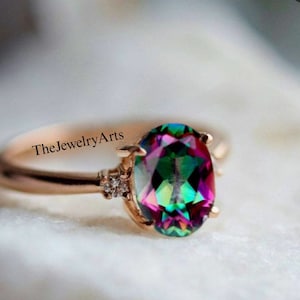 Pode incluir: Um anel de ouro rosa com uma grande gema oval iridiscente e gemas menores transparentes na banda. O texto "TheJewelryArts" é visível na banda.