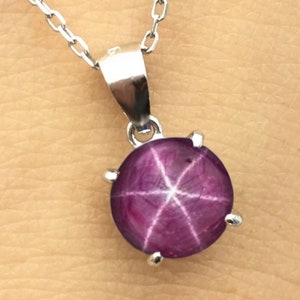 Puede incluir: Un collar de cadena de plata con un colgante que presenta una piedra preciosa de rubí estrellado redonda de color rosa oscuro.
