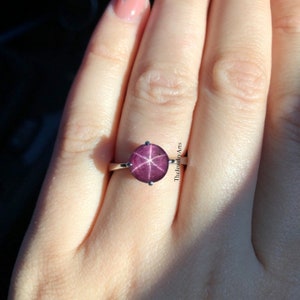 May include: A silver ring with a round, dark pink star ruby gemstone. The ruby has a star-shaped pattern inside.