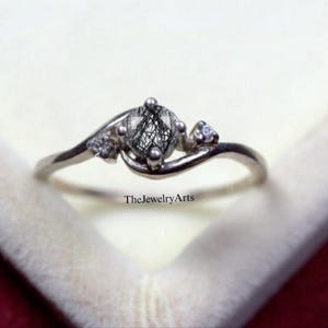 May include: A silver ring featuring a central, round, black and grey stone, with two smaller clear stones either side. The ring's band curves, with the text "TheJewelryArts" visible.