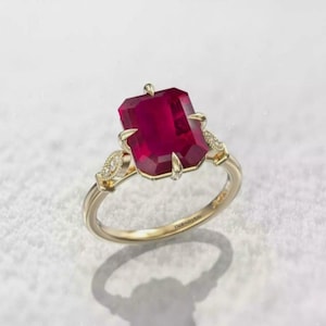 May include: A gold ring featuring a large, rectangular, deep red gemstone. The ring has intricate detailing with smaller accent stones and a delicate band. The Jewelry Arts is engraved on the band.