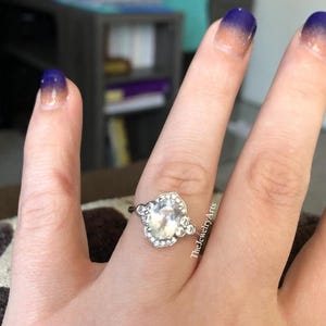May include: A silver ring with a large, clear, oval-shaped gemstone, surrounded by smaller, clear stones. The ring is on a finger with blue and orange ombre nail polish. The Jewelry Arts is written on the side of the finger.