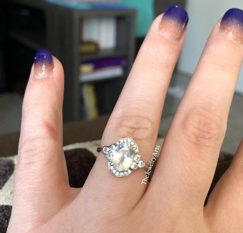 May include: A silver ring with a large, clear, oval-shaped gemstone, surrounded by smaller, clear stones. The ring is on a finger with blue and orange ombre nail polish. The Jewelry Arts is written on the side of the finger.
