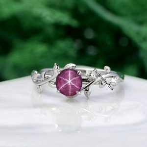 Puede incluir: Un anillo de plata con un delicado diseño de vid que presenta hojas y una piedra preciosa de rubí estrellado rosa redonda. El texto "The Jewelry Arts" es visible en el anillo.