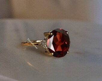 May include: A gold ring with a large, round, red gemstone surrounded by smaller, clear gemstones. The ring is on a white surface.