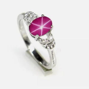 Op de afbeelding: Een zilveren ring met een grote, ovaal geslepen ster-robijn edelsteen. De robijn vertoont een zesstralig ster-effect. De ring heeft een slanke band en is versierd met kleinere, heldere edelstenen. De tekst "TheJewelry Arts" is zichtbaar op de band.