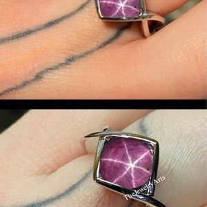 May include: A silver ring with a pink star-shaped gemstone set in a square setting. The ring is on a finger and the gemstone has a starburst pattern.