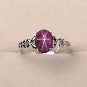 Può includere: Un anello d'argento con una pietra preziosa di rubino stellato rosa. L'anello ha un delicato design floreale.