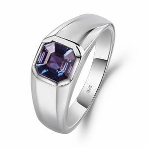 May include: A polished silver ring featuring a square-cut, multifaceted purple and blue gemstone set in the center. The band is smooth and wide, with the hallmark 925 stamped clearly on the inner surface, indicating sterling silver construction.