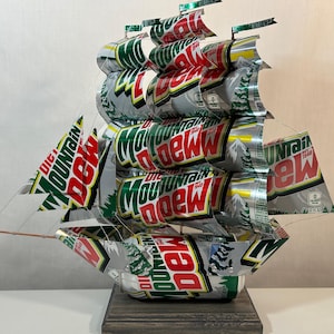 May include: A handcrafted ship sculpture made from recycled Mountain Dew soda cans. The sails and hull are constructed from the cans, showcasing the brand's logo in green, red, and white. The ship is mounted on a dark wooden base, creating a unique decorative piece.