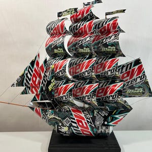 May include: A handcrafted ship sculpture made from repurposed beverage cans. The sails and hull feature bold red, black, and white graphics with the text "MTN DEW Zero Sugar Baja Blast". The ship is mounted on a black base.