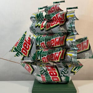 May include: A handcrafted ship sculpture made from recycled Diet Mountain Dew cans. The sails and hull are constructed from the cans, with the "Mountain Dew" logo visible. The ship is mounted on a green wooden base.