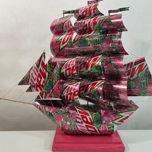 May include: A handcrafted ship sculpture made from recycled Mountain Dew soda cans. The ship features red, white, and green colors, with the Mountain Dew logo visible on the sails and hull. The ship is mounted on a pink wooden base.
