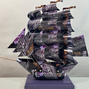 May include: A handcrafted ship sculpture made from recycled Citrus Blackcherry Game Fuel beverage cans. The sails and hull are constructed from the cans, featuring purple and black graphics. The ship rests on a purple wooden base, with small flags at the top of the masts.
