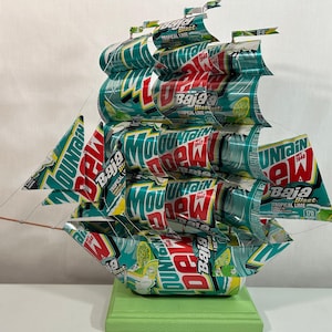 May include: A handcrafted ship sculpture made from recycled Mountain Dew Baja Blast cans. The ship features multiple sails and a green base. The cans display the Mountain Dew logo and tropical lime graphics.