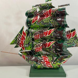 May include: A handcrafted ship sculpture made from recycled Mountain Dew Thrashed Apple cans. The sails and hull are constructed from the green, red, and white cans, mounted on a green base. The sculpture features the Mountain Dew logo and "Thrashed Apple" text.