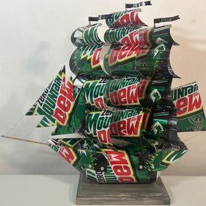 May include: A handcrafted ship sculpture made from recycled Mountain Dew soda cans. The ship features green, red, and white colors, with the Mountain Dew logo visible on the sails and hull. The sculpture is mounted on a wooden base.