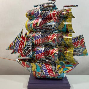May include: A handcrafted ship sculpture made from recycled Mountain Dew Spark soda cans. The ship features multiple sails and a hull constructed from the colorful cans, with the "Spark" logo visible. The sculpture is mounted on a purple wooden base.