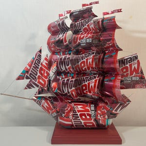 May include: A handcrafted ship sculpture made from red and white Mountain Dew Code Red Cherry soda cans. The sails and hull are constructed from the cans, with the words "Mountain Dew" and "Code Red Cherry" visible. The ship sits on a red wooden base.