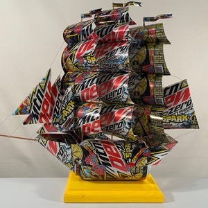 May include: A ship model made entirely of Mountain Dew Zero Sugar cans. The ship has a yellow base and is decorated with red, white, and blue details. The ship's sails are made of the cans, and the ship's hull is made of the cans.