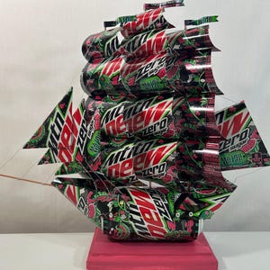 May include: A handcrafted ship sculpture made from recycled Mountain Dew Zero Sugar cans. The ship features red, green, and black colors, with the brand's logo visible on the sails and hull. The sculpture is mounted on a pink wooden base.