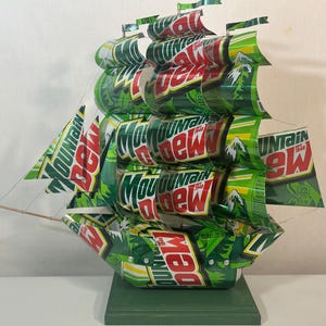 May include: A handcrafted ship sculpture made from recycled Mountain Dew soda cans. The green, white, and red colors of the cans are prominently displayed, with the "Mountain Dew" logo visible on the sails and hull. The ship sits on a green wooden base.