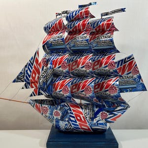 May include: A handcrafted ship sculpture made from repurposed Mountain Dew Voltage cans. The sails and hull are constructed from the red, white, and blue printed aluminum cans, mounted on a blue wooden base. The ship features the text "Voltage".