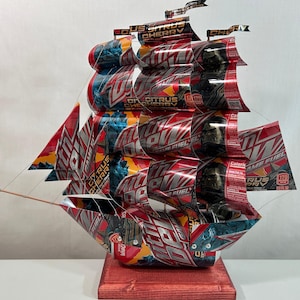 May include: A handcrafted ship sculpture made from recycled aluminum beverage cans. The ship features red, white, and blue colors, with the words "Citrus Cherry" and "Game Fuel" visible on the sails and hull. It is mounted on a stained wooden base.