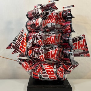 May include: A handcrafted ship sculpture made from Zero Sugar Mountain Dew Code Red cherry soda cans. The red and black ship features multiple sails and is mounted on a black base. The sails display the Mountain Dew logo.
