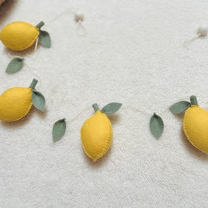 May include: A decorative garland featuring four yellow felt lemons with green felt leaves attached to a thin white string. The lemons are evenly spaced along the cord, which also includes small white spherical beads between the fruit elements.