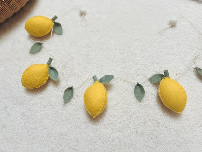 May include: A decorative garland featuring four yellow felt lemons with green felt leaves attached to a thin white string. The lemons are evenly spaced along the cord, which also includes small white spherical beads between the fruit elements.