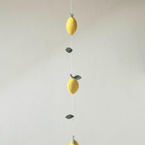 May include: A vertical hanging garland featuring three yellow felt lemons and several small green felt leaves attached to a thin white string. The lemons are evenly spaced along the cord, each topped with a small green stem and leaf detail.