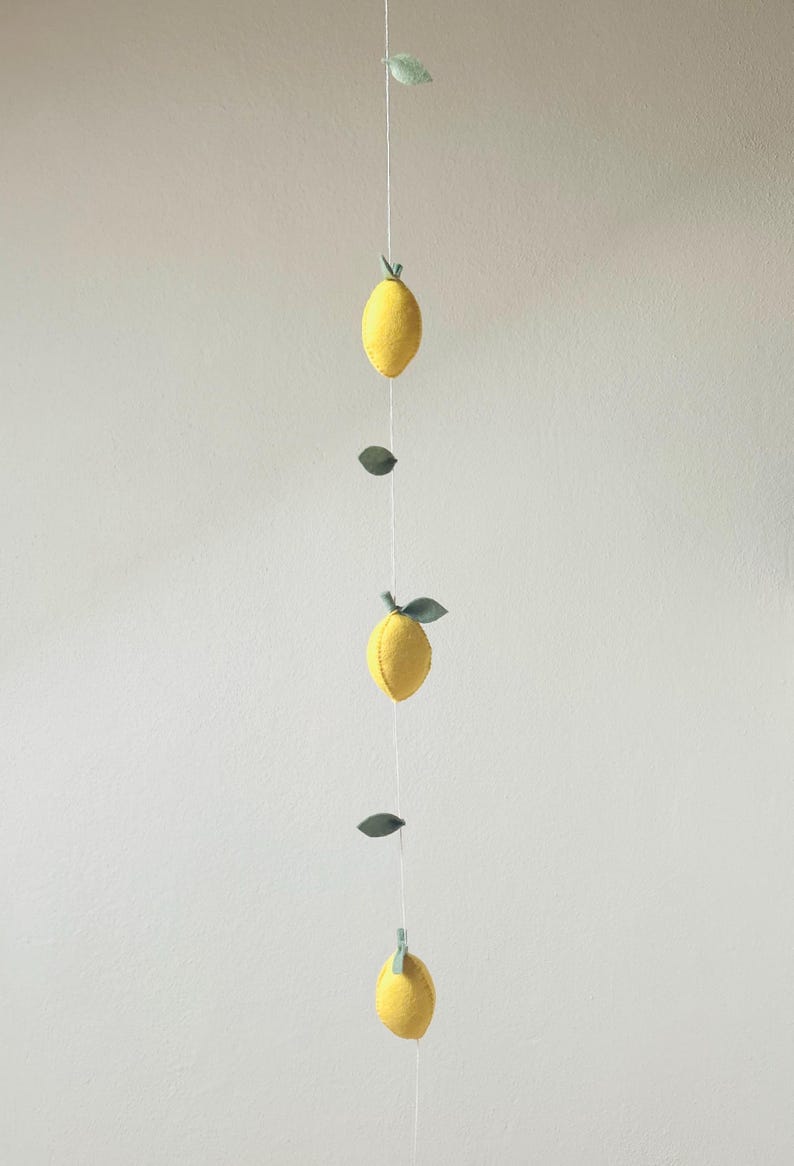May include: A vertical hanging garland featuring three yellow felt lemons and several small green felt leaves attached to a thin white string. The lemons are evenly spaced along the cord, each topped with a small green stem and leaf detail.