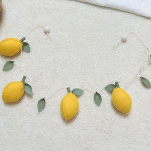May include: A decorative garland featuring five stuffed yellow lemon shapes made of felt, each accented with small sage green felt leaves. The lemons are strung together on a thin white cord, with small wooden beads at the ends for hanging.
