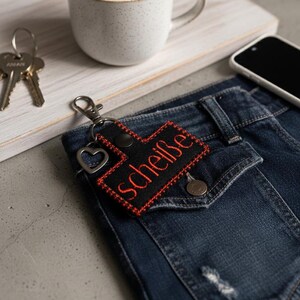 Könnte beinhalten: Schwarzer Schlüsselanhänger mit dem Wort "scheiße" in Rot gestickt, an einer blauen Jeans befestigt. Der Schlüsselanhänger hat einen herzförmigen Anhänger. Eine Tasse Kaffee und ein Smartphone sind ebenfalls sichtbar.