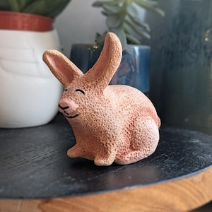 May include: A ceramic rabbit ornament in peach and pink hues, featuring a textured finish. The rabbit has long ears, a pink nose, and a smiling face. It rests on a dark surface, with blurred background elements visible.