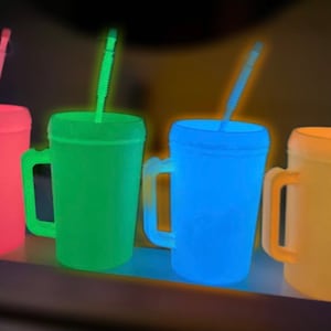 Op de afbeelding: Vier glow-in-the-dark plastic bekers met handvatten en rietjes. De bekers zijn roze, groen, blauw en geel.